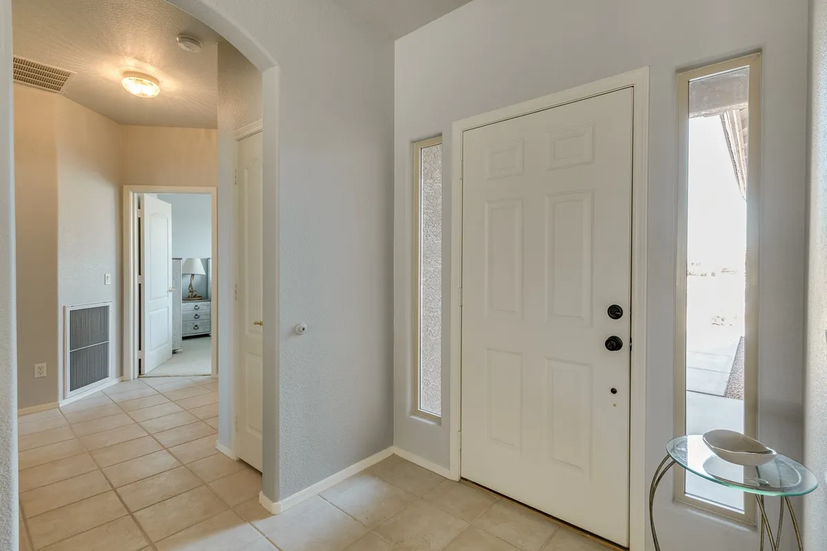 This image shows a welcoming front entryway with a bright front door framed by side windows. A hallway leads to other rooms, and a small table adds a personal touch to the space. The scene is inviting, offering a sense of arrival and transition into the home.