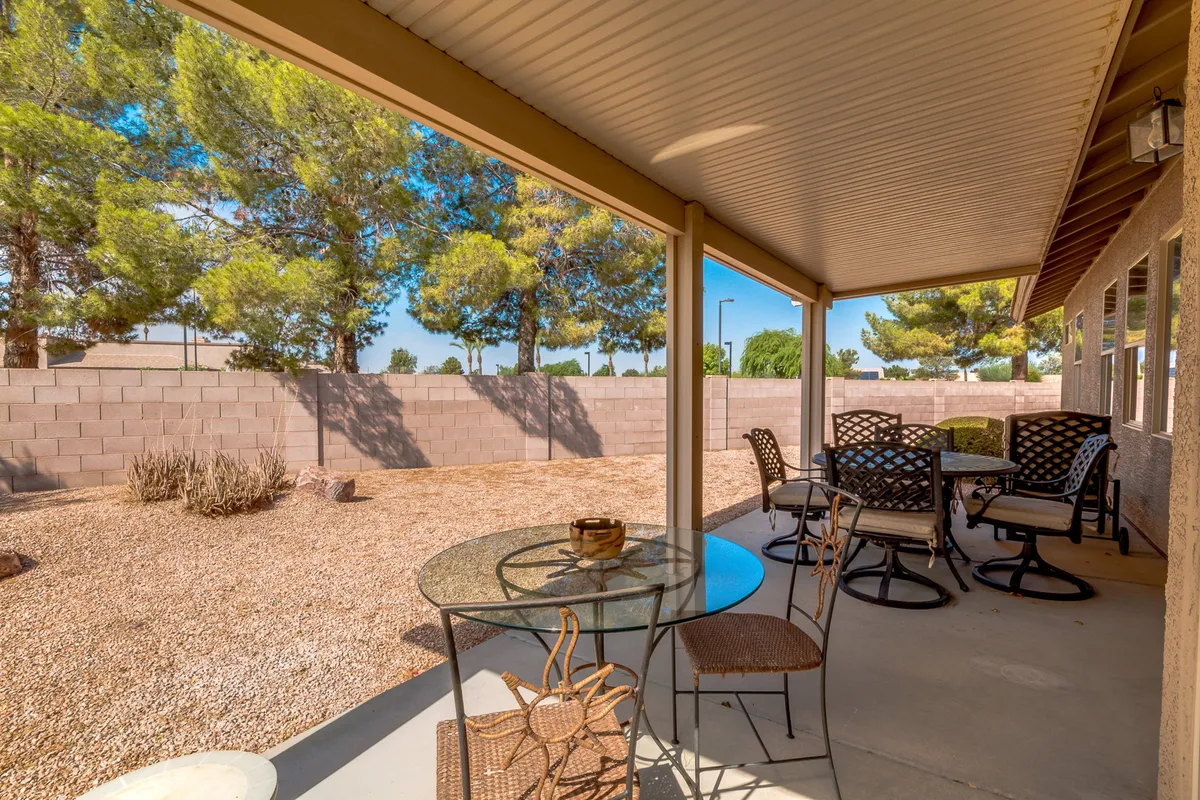 A peaceful patio scene with a round table and comfortable swivel chairs set on a landscaped gravel area. Surrounded by trees and desert-friendly plants, the setting showcases the easy-care landscaping and the flexibility for both casual meals and special moments outdoors.