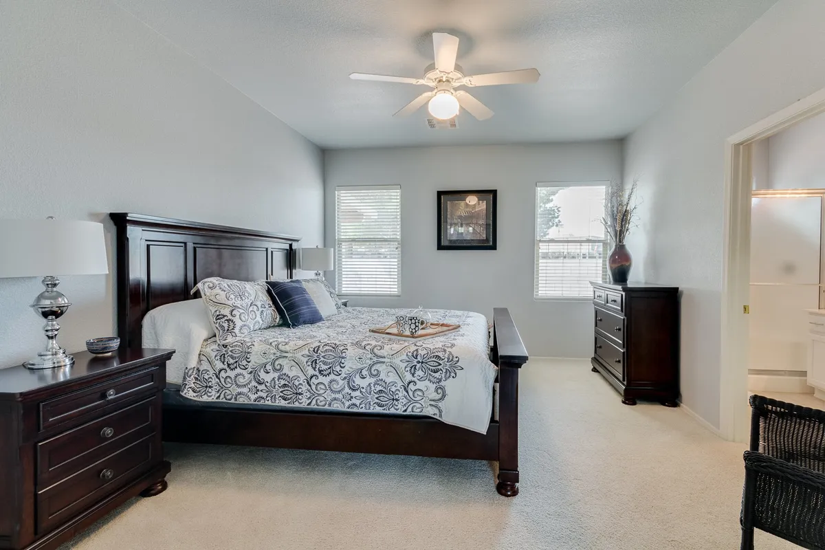 A king-sized bed with an ornate wooden frame takes center stage in this spacious, serene master suite. Decorative pillows, coordinated bedding, and elegant artwork create a tranquil atmosphere. Nightstands with matching lamps flank the bed, while ample natural light pours in through a generous window overlooking the backyard.