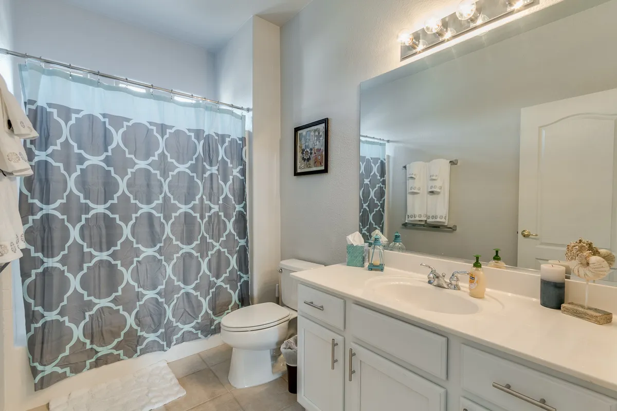 A well-lit bathroom with a vanity featuring double sinks and a large mirror. Decorative details like the patterned shower curtain and thoughtfully arranged accessories add a homey touch. The space is bright and inviting, ideal for unwinding after a long day.