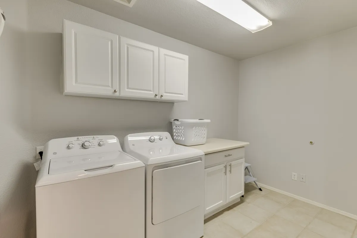 A bright, functional laundry room featuring a modern washing machine and dryer, white overhead cabinets for organized storage, and a laundry basket placed conveniently for use. The space is clean and thoughtfully arranged, emphasizing practicality and ease of use.