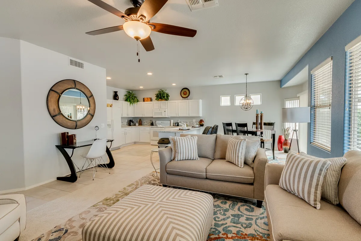 This image captures the heart of the open-concept great room, featuring a comfortable sofa, a striped ottoman, a dining table, a glimpse into the kitchen area, and a decorative mirror. The space radiates warmth and sociability, with modern furnishings and plenty of natural light that underscores the fresh, airy ambiance described in the section.