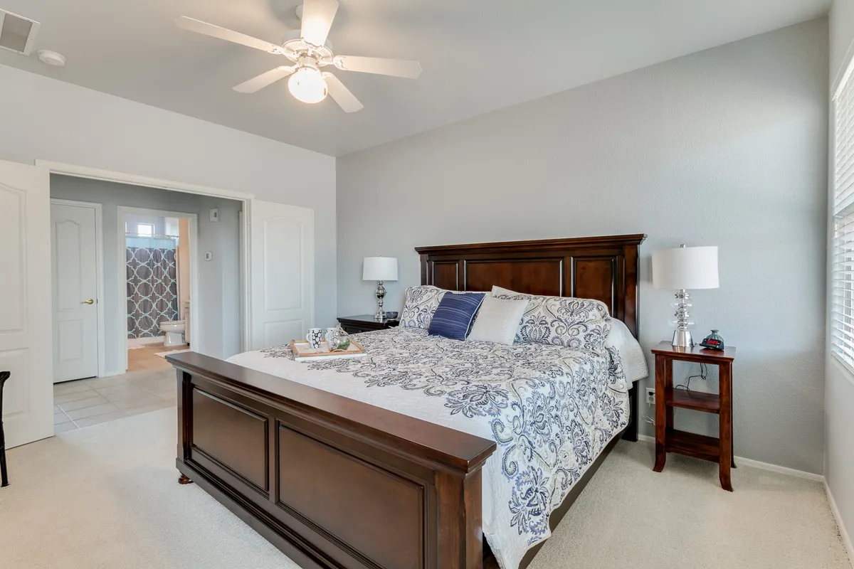 A cozy guest bedroom with a comfortable bed dressed in decorative bedding. A nightstand with a lamp sits beside the bed, and an open doorway reveals the adjoining bathroom, emphasizing convenience for guests or family.